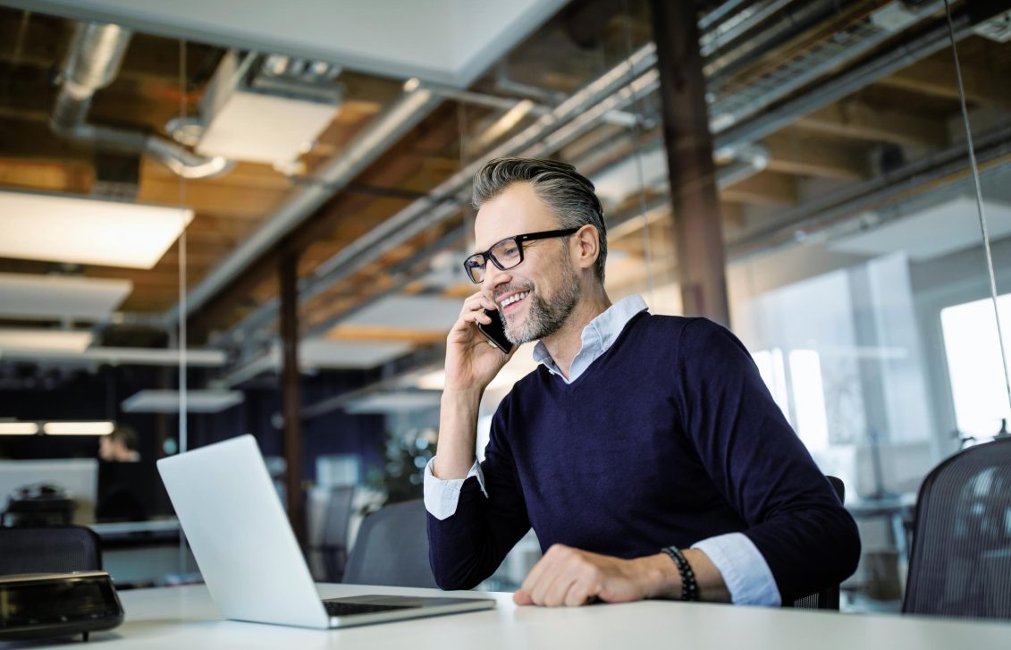 Motorrijtuigen schadebehandelaar bij Baloise aan het werk. Hij zit achter zijn bureau met laptop en voert een telefoongesprek.