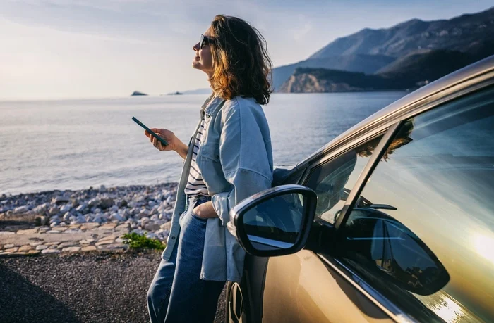 Foto van een vrouw met zonnebril en telefoon in haar hand, die leunend tegen haar auto geniet van de laatste zonnestralen aan zee.