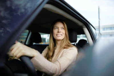 Foto van een jonge vrouw achter het stuur van haar auto, die glimlachend door haar zijruit naar buiten kijkt.