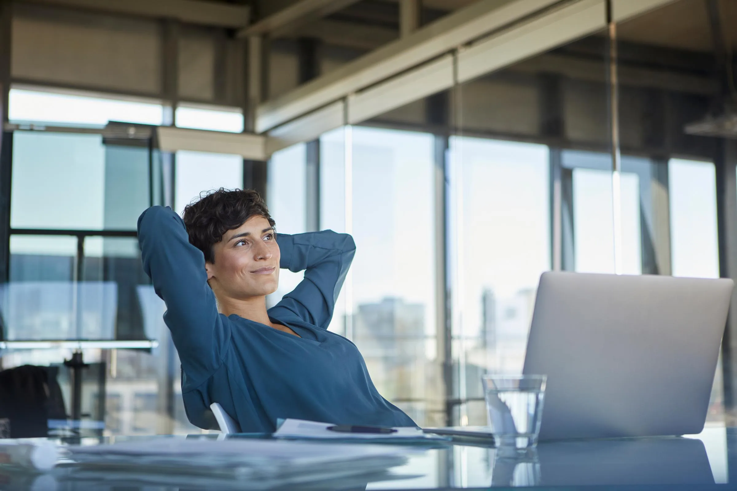 Foto van een tevreden in de verte kijkende vrouw met armen in de nek zittend aan haar bureau met laptop op kantoor.
