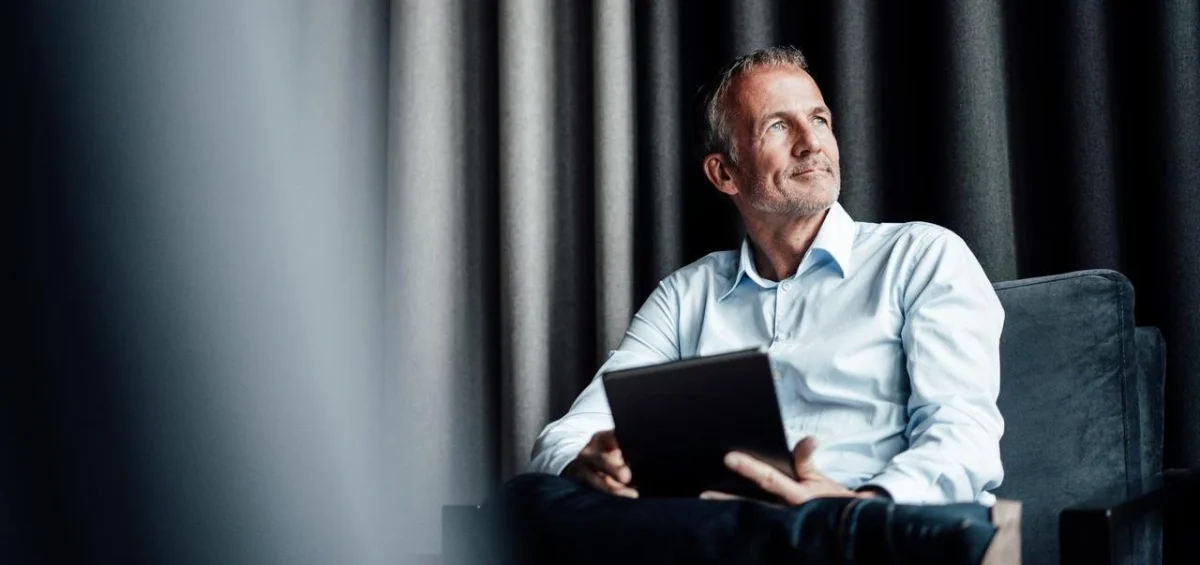 Foto van een zakenman met tablet die wegkijkt terwijl hij in een fauteuil zit te wachten in de lobby van een conferentiecentrum.