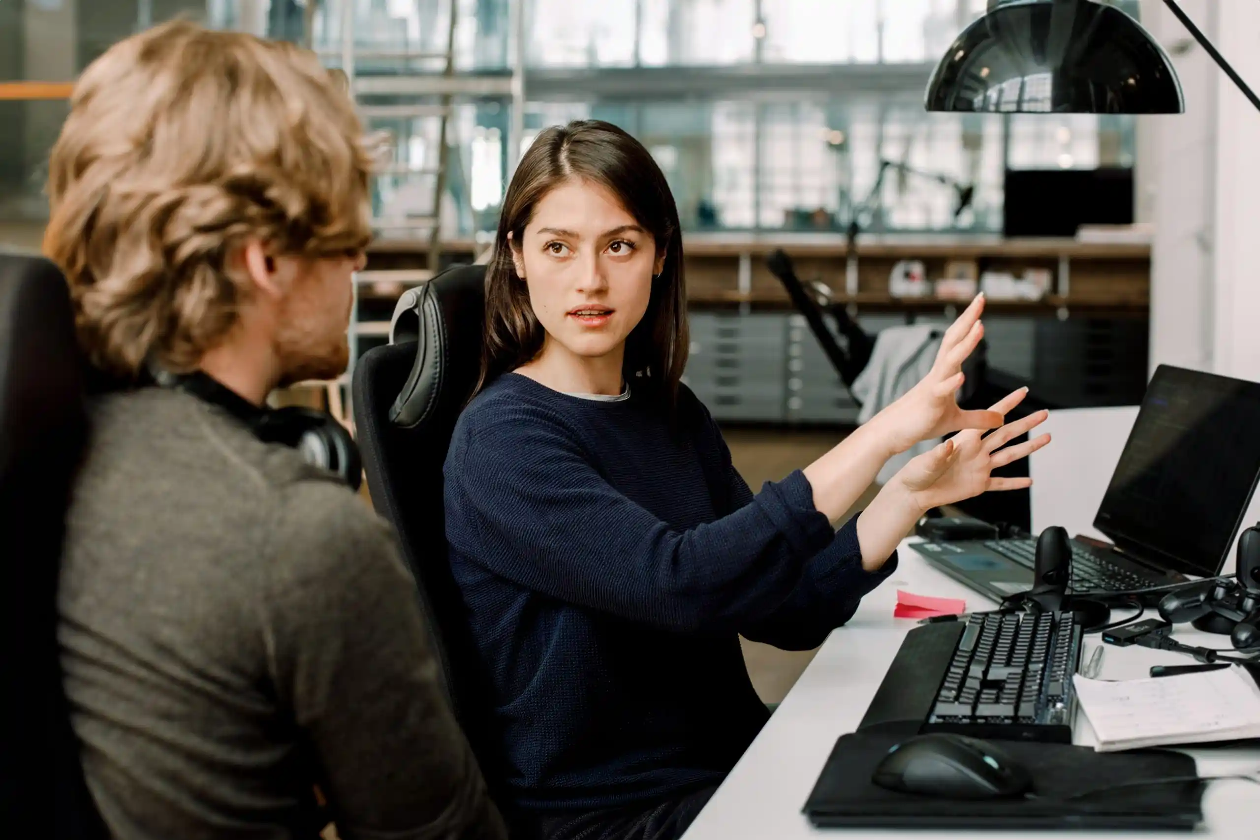 Foto van man die aandachtig luistert naar een vrouwelijke collega die, al gebarend met haar handen, iets aan het uitleggen is aan haar bureau op kantoor.