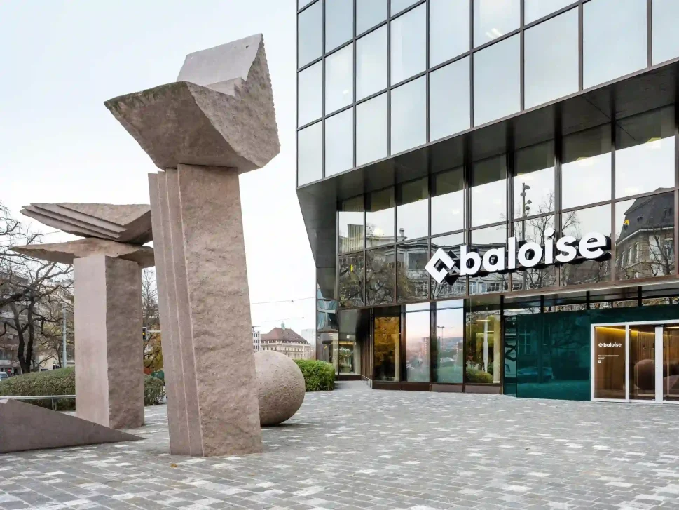Foto van de entree van het hoofdkantoor van Baloise in Bazel, Zwitserland met glazen gevel en abstracte stenen sculptuur voor het pand.