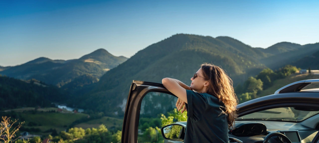 Foto van een vrouw met zonnebril die leunend op haar bestuurdersportier van haar auto aan het genieten is van de laatste zonnestralen van die dag in de bergen.