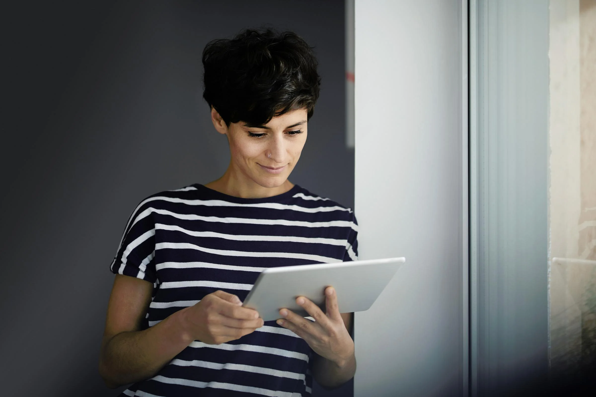 Foto van een jonge vrouw met kort donker haar die bij het raam naar haar tablet staat te kijken.