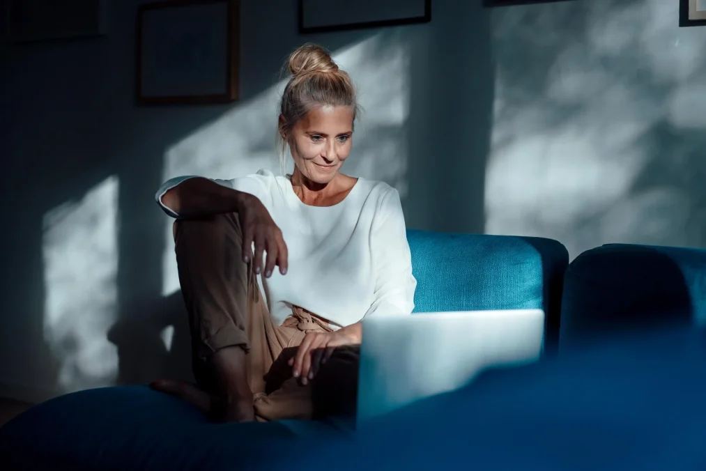 Foto van een vrouw die in een relaxte zit op de bank tevreden naar haar laptop kijkt.