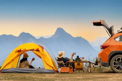 Foto van een auto waar de achterklep van open staat, een vrouw in een klein tentje en een man in de campingstoeltje voor de tent die genieten van het uitzicht in de bergen.