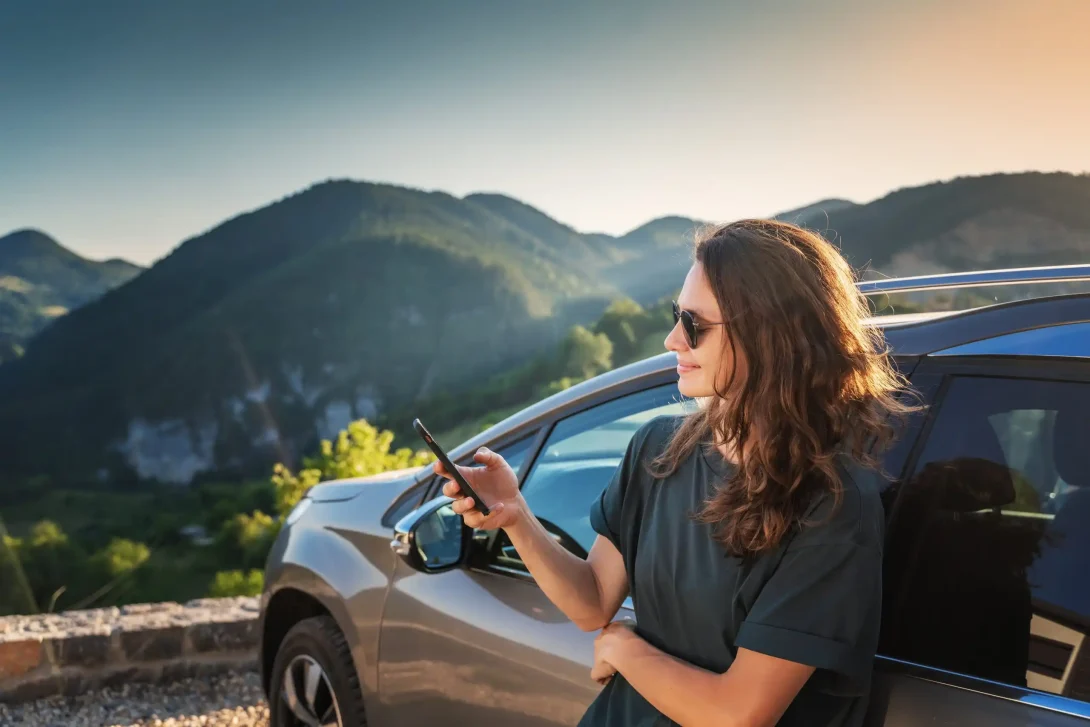 Foto van een vrouw met zonnebril die tegen haar auto aan leunt terwijl ze op haar telefoon kijkt tijdens de zonondergang.