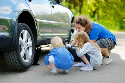 Foto van een vader die bezig is zijn auto op te krikken om de achterband te verwisselen terwijl zijn 2 jonge dochters aandachtig toekijken.