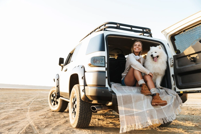 Foto van een jonge vrouw en haar hond (samojeed) die lachend vanuit de achterklep van haar terreinwagen naar de zandvlakte kijkt.