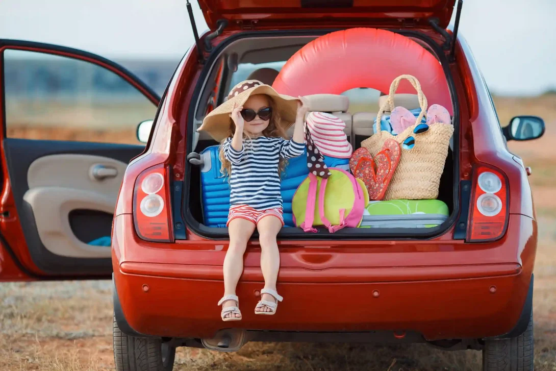 Foto van een jong meisje met grote strohoed die in de achterbak van een auto zit die vol zit met bagage voor een zonvakantie.