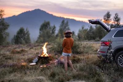Foto van een vrouw die een foto maakt van een picknick met kampvuur in de wildernis. De auto met openstaande klep waar alles in uitgekomen is, is ook nog te zien op de foto.