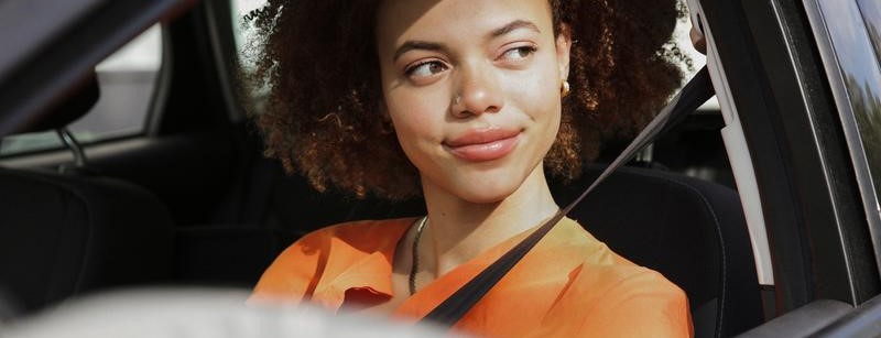 Foto van een jonge vrouw achter het stuur van haar auto, die zelfverzekerd door haar zijruit naar buiten kijkt.