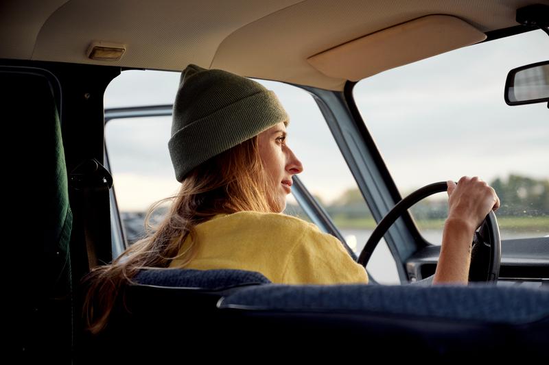 Foto van een jonge vrouw met muts die achter het stuur van haar auto naar de zonsondergang kijkt.