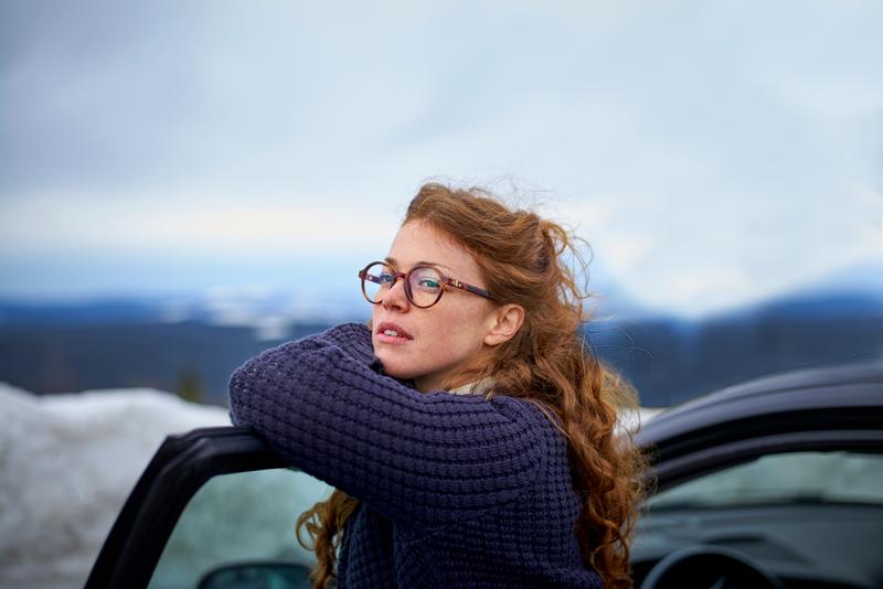 Foto van een vrouw die op haar autodeur leunt en uitkijkt over het natuurlandschap terwijl ze even pauzeert tijdens het rijden.