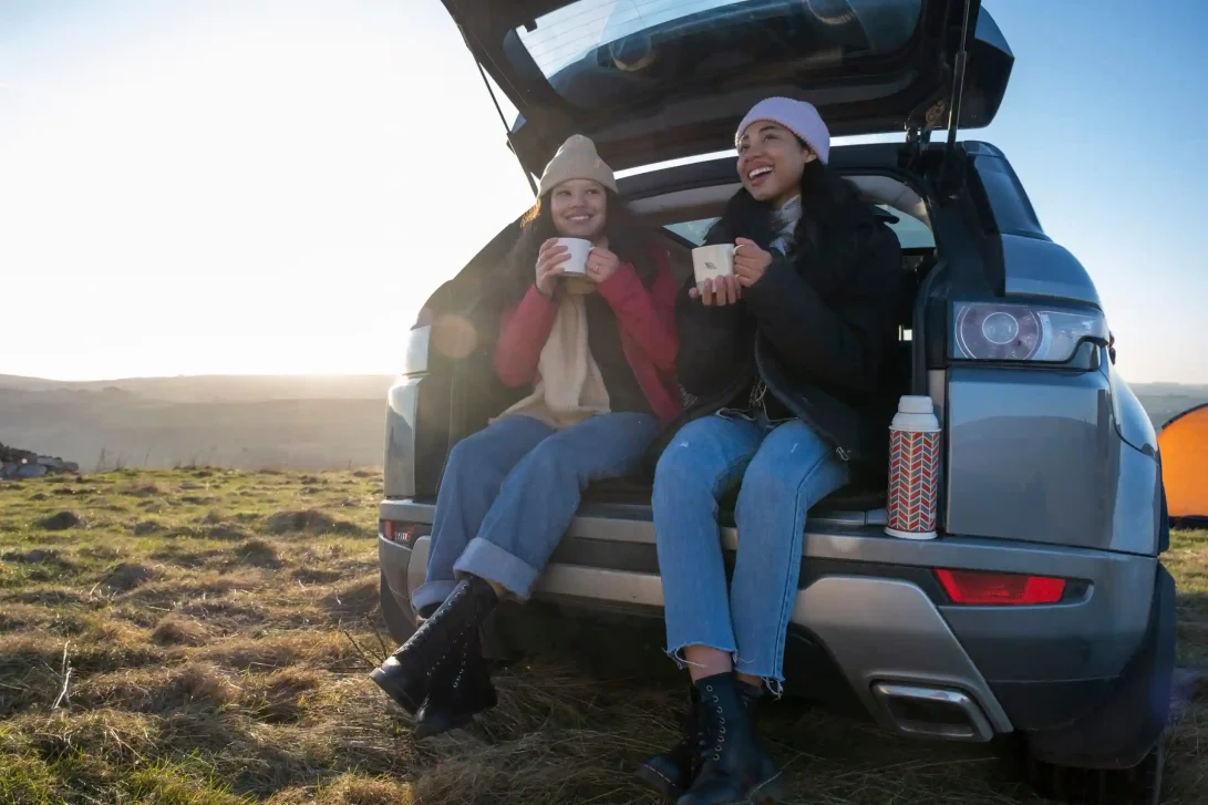 Foto van 2 vriendinnen met mutsen op die uit een mok drinken vanuit de kofferbak van een auto in de schotse hooglanden.