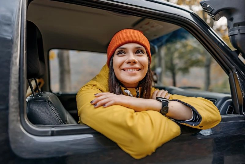 Foto van een vrouw met muts die lachend naar buiten kijkt door het open raam aan de passagierskant van de auto.