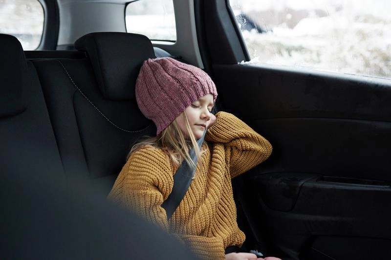 Foto van een klein meisje met dikke trui en muts die aan het slapen is op de achterbank van een auto die door een besneeuwd landschap rijdt.