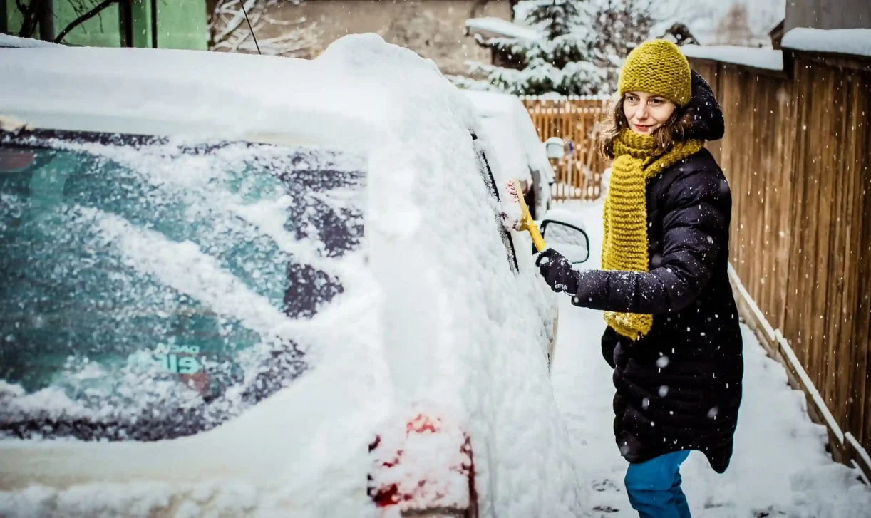 Foto van een vrouw die met een borstel haar auto sneeuwvrij aan het maken is.