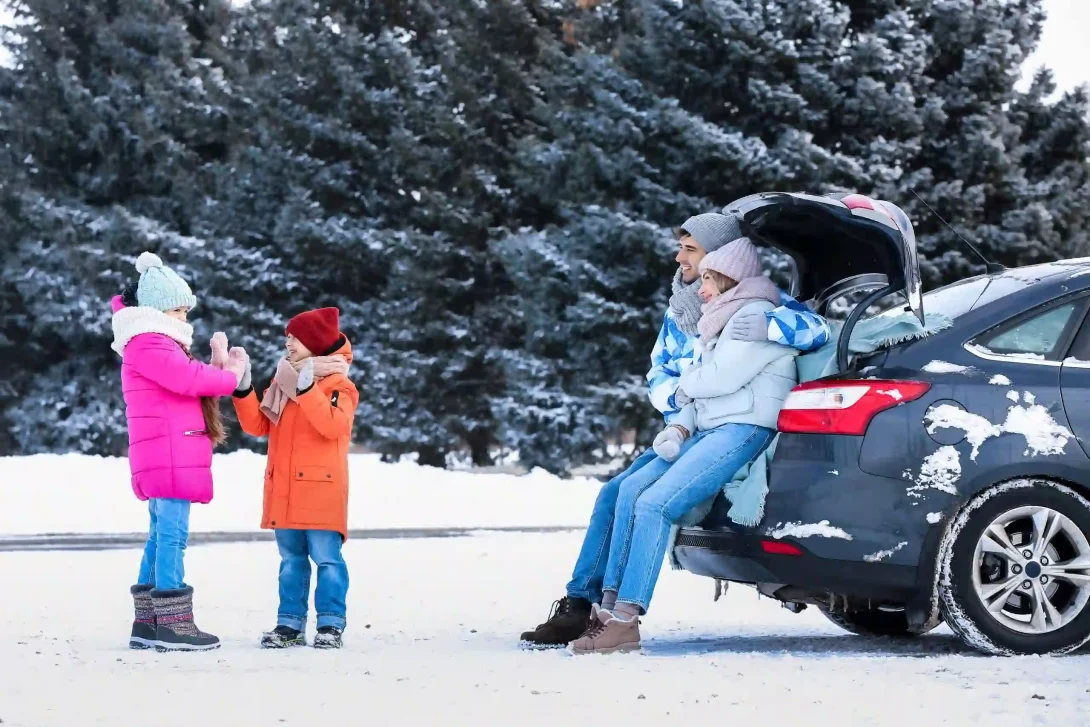 Foto van 2 ouders die vanuit de kofferbak van hun auto naar hun spelende kinderen kijken op een besneeuwde winterdag.