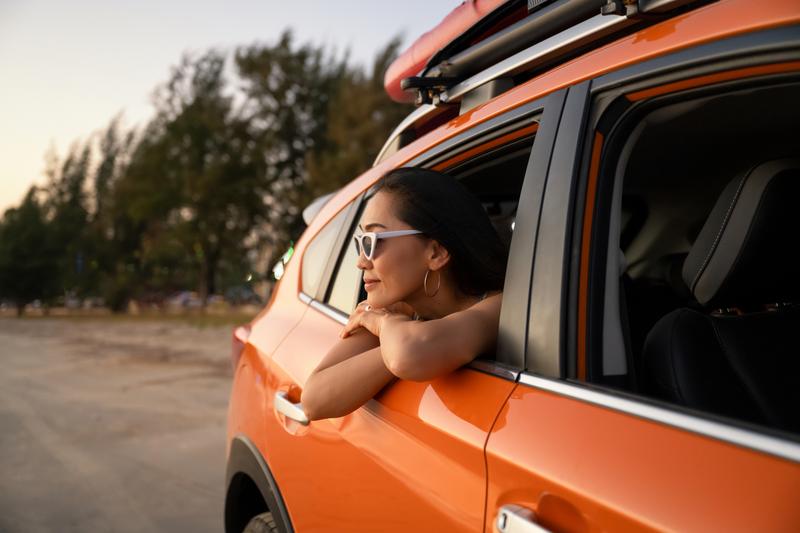 Foto van een vrouw met witte zonnebril die tevreden vanuit het open achterraam van een auto naar de zonsondergang kijkt.