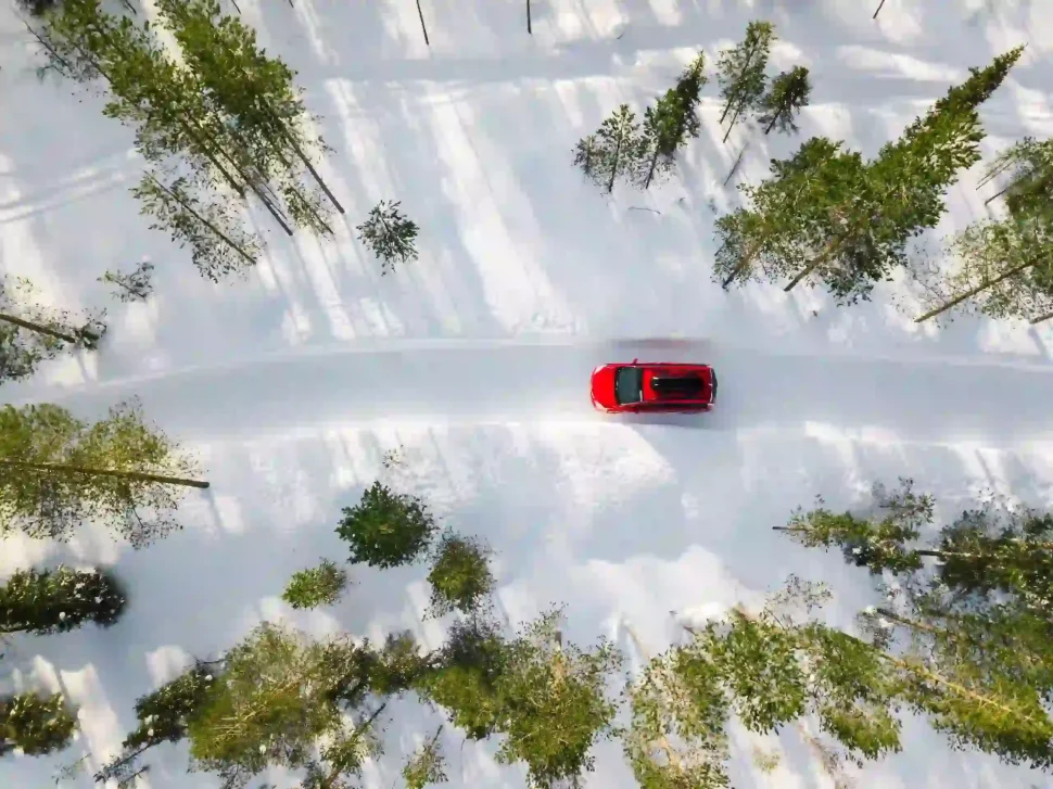 Luchtfoto van een rode auto met een zwarte dakkoffer die door het witte sneeuwlandschap van een winterbos rijdt op een landweg in Finland, Lapland.