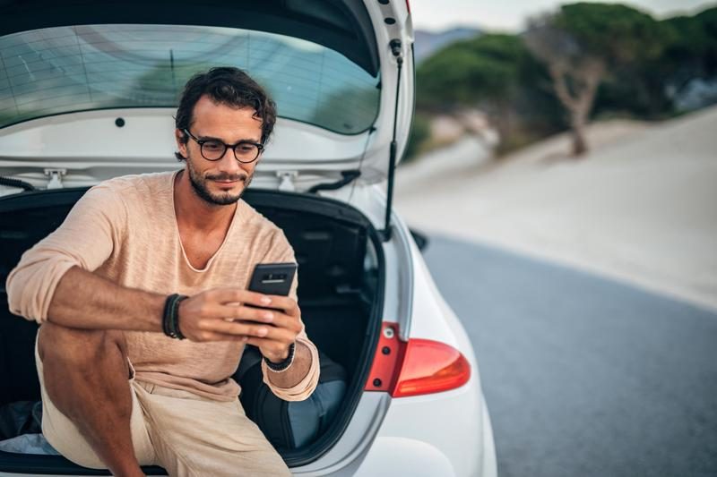 Foto van een man die op het einde van een zomerse dag op zijn telefoon kijkt terwijl hij ontspannen in de achterklep van zijn auto zit.