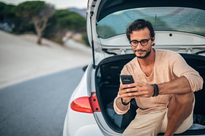 Foto van een man die op het einde van een zomerse dag op zijn telefoon kijkt terwijl hij ontspannen in de achterklep van zijn auto zit.