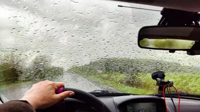 Foto waarbij we vanuit de berijdersstoel van een auto naar buiten kijken door de voorruit vol met regendruppels terwijl we aan het rijden zijn.