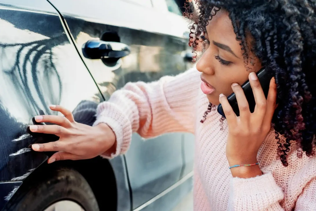 Foto van een zwarte jonge vrouw die de schade aan het linker voorspatbord van haar auto bekijkt en belt tegelijkertijd haar verzekeringsmaatschappij.